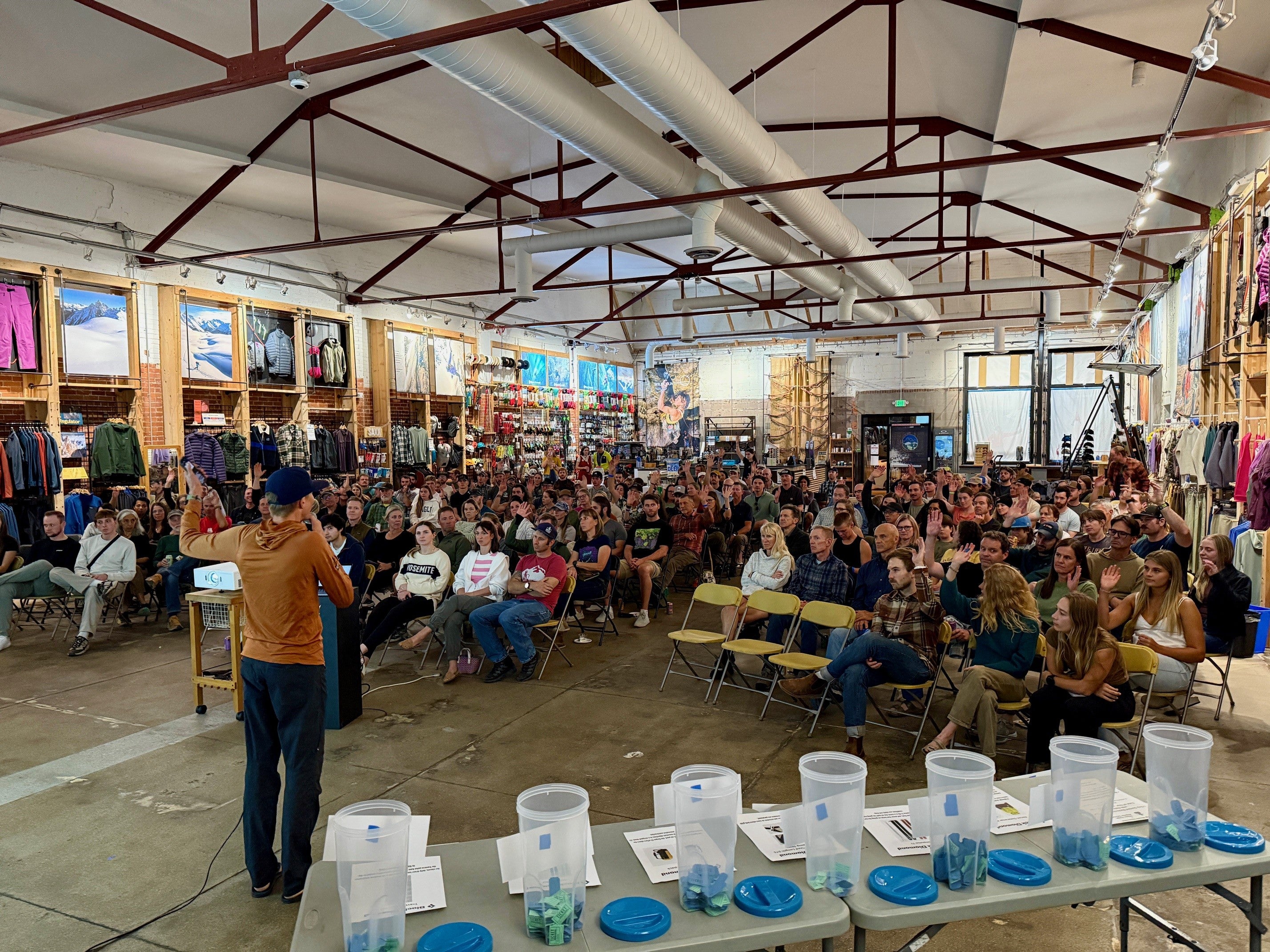 Large indoor event with a speaker addressing an audience in a warehouse-like setting.